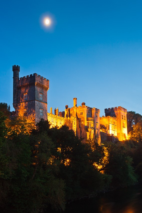 Photo of the moonrise over Lismore Castle in County Waterford, Ireland with the castle being illuminated by yellow lights