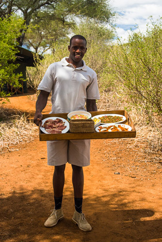Photo of a smiling african man offering various foods on a tray in a safari camp in Tansania, East Africa